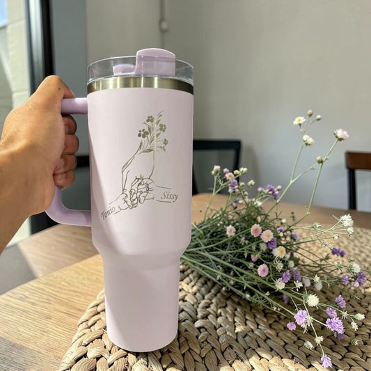 Pink tumbler with floral design held by a hand, placed on a woven mat with flowers.
