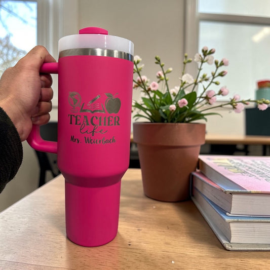 Pink travel mug with 'Teacher Life' text held by a hand on a desk with books and a plant.