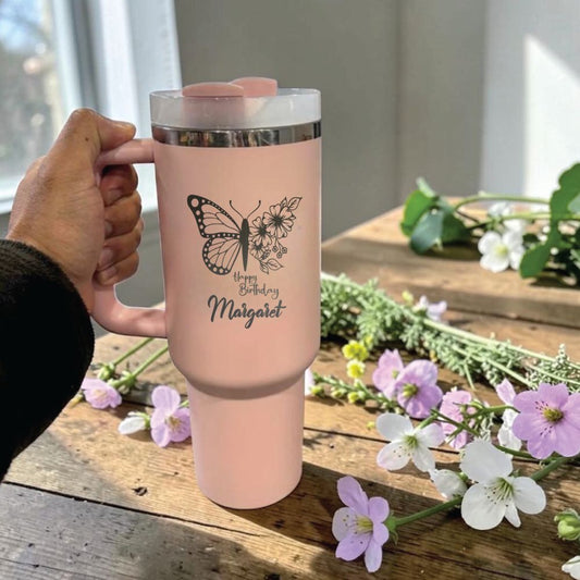 Pink Stanley 40 0z tumbler for birthday gift with butterfly design and text held by a hand on a wooden surface with flowers.
