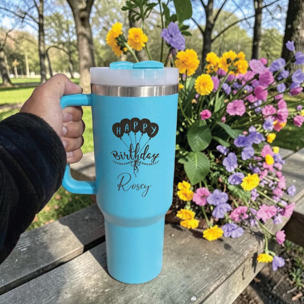 Blue Stanley 40 0z tumbler for birthday  with personalized text held outdoors next to a flower arrangement