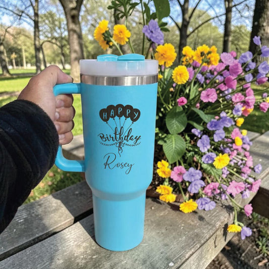 Blue Stanley 40 0z tumbler for birthday  with personalized text held outdoors next to a flower arrangement