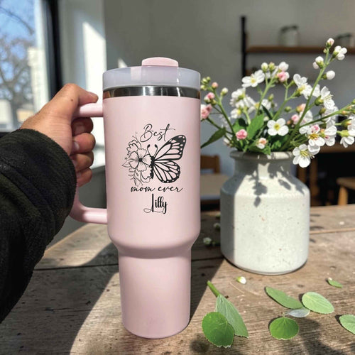 Pink travel mug with butterfly design held by a hand on a wooden table with flowers in the background