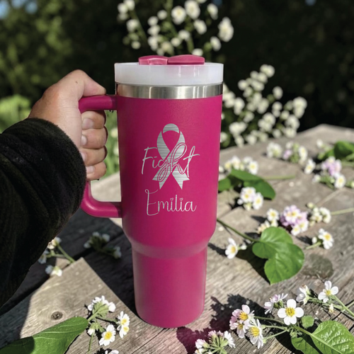 Pink Stanley 40 0z tumbler for cancer awareness with 'Fight Emilia' design held by a hand on a wooden surface with flowers.