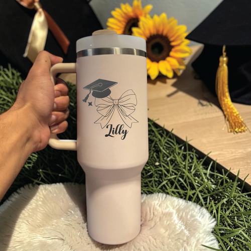 White travel mug with graduation cap and bow design, held by a hand, sunflower and tassel in the background.