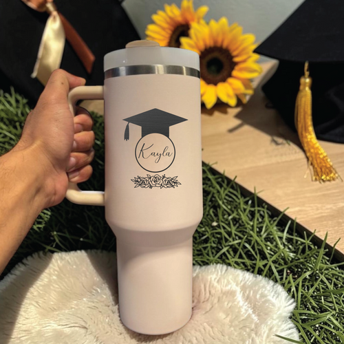Graduation-themed Stanly 40 0z tumbler with a design and name, held by a hand with sunflowers and a cap in the background.