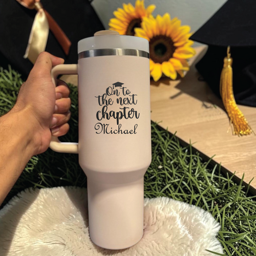 White travel  Stanley 40 0z tumbler with 'On to the next chapter Michael' text, held by a hand, with sunflowers and a graduation cap in the background.