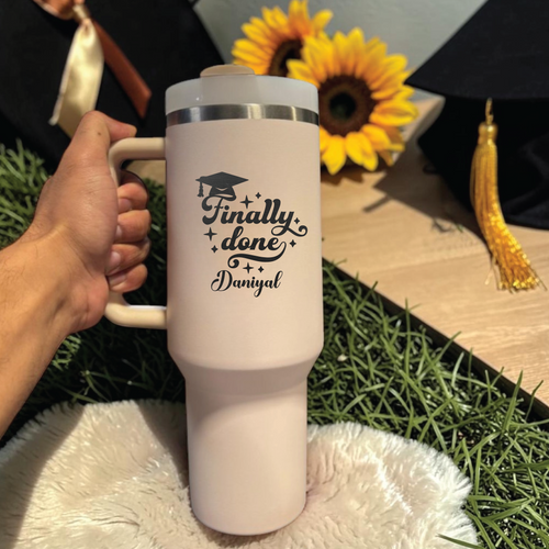 Person holding a white mug with 'Finally done Daniyal' text, sunflowers, and graduation cap in the background.