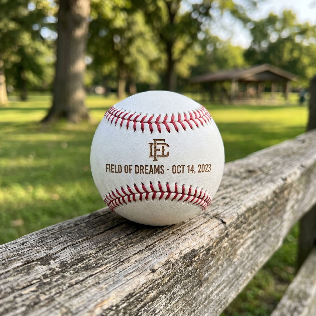 A white baseball resting on a weathered wooden fence in a park, featuring red stitching and a custom printed design with the letters 'FD' and the text 'FIELD OF DREAMS - OCT 14, 2023'.