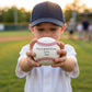 A young boy wearing a navy baseball cap and white shirt stands on a baseball field holding a white baseball with red stitching and printed text that reads 'I'm so proud of you, Love, Dad'.