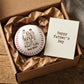 A personalized baseball-shaped keepsake or token with a family illustration and the words 'Dad MVP' sits in a brown gift box with crumpled tissue paper, next to a card that reads 'Happy Father's Day'.
